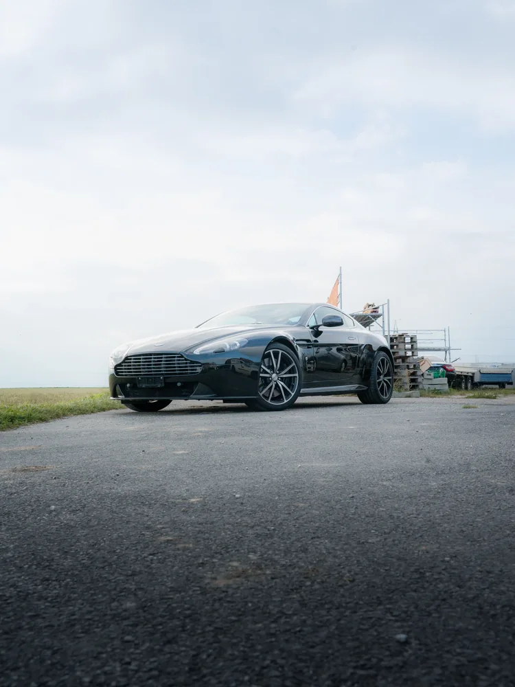 Aston Martin Vantage S noire en covering partiel, de trois quarts avant, ciel voilé